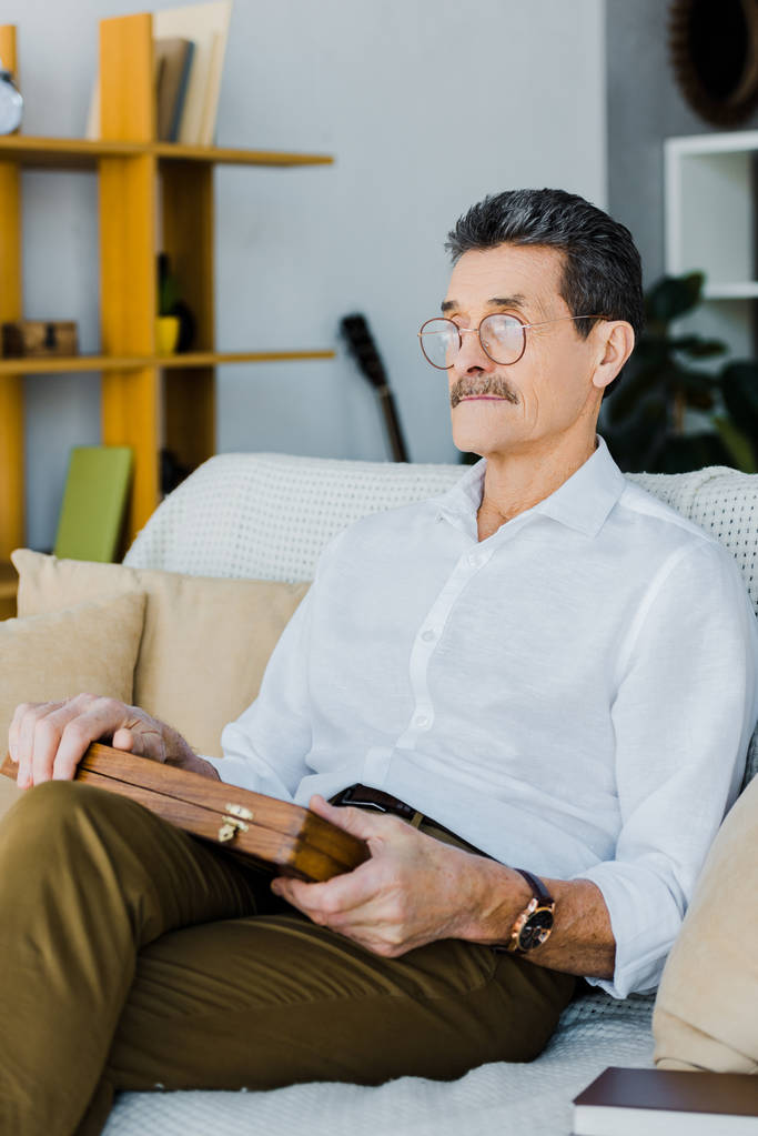 důchodce v brýlích drží šachovnici při sezení na pohovce - Fotografie, Obrázek