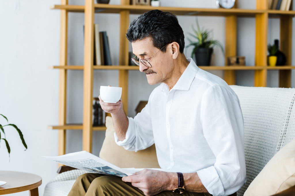 aposentado homem de óculos segurando xícara com café e lendo jornal em casa
 - Foto, Imagem