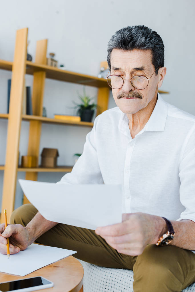 hombre mayor en gafas sosteniendo lápiz y mirando el papel en casa
 - Foto, imagen