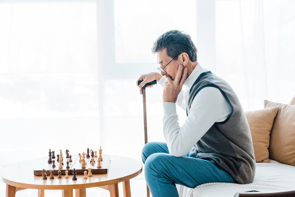 Nachdenklicher Rentner mit Brille blickt auf Schachbrett und hält Gehstock in der Hand  - Foto, Bild