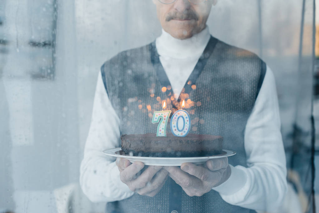 обрезанный вид грустного пожилого человека, держащего торт на день рождения у окна
 - Фото, изображение