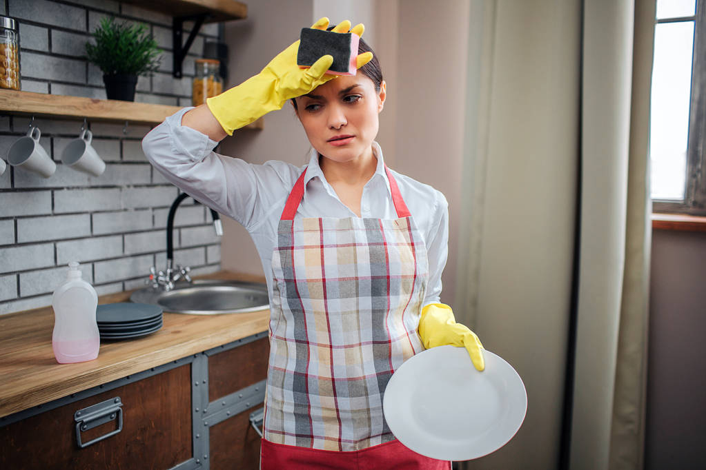 Tired female cleaner stand in kitchen and hold hand on forehead. She wear apron and yellow gloves. Woman hold white plate. - Photo, Image