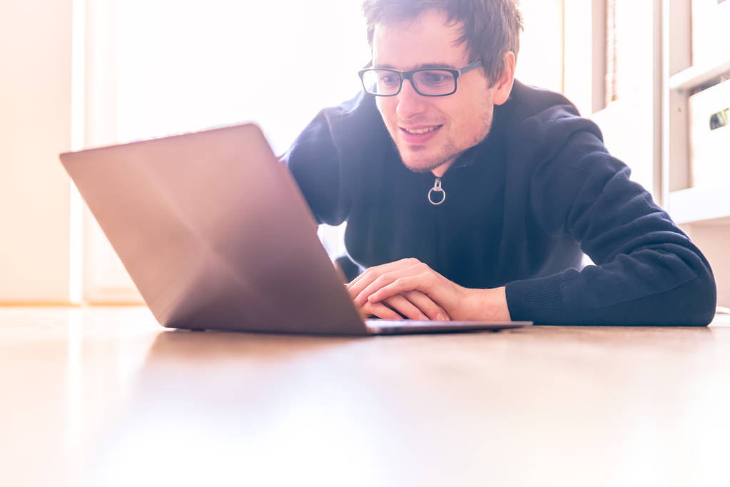 幸せノート パソコンで若い男が木の床に取り組んでいる、光が差し込む窓から - 写真・画像