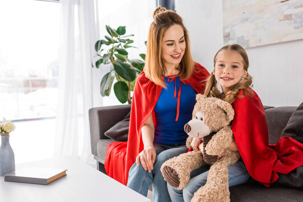 Gyönyörű anya és a gyerek a piros köpeny gazdaság mackó, és ül a kanapén, otthon - Fotó, kép