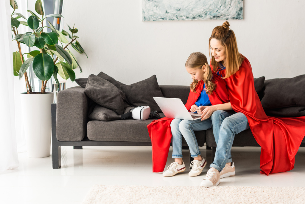 mother and daughter in red cloaks sitting on sofa and holding laptop - Photo, Image