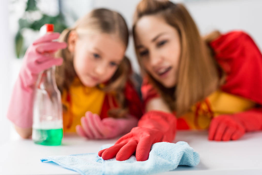 Селективное внимание матери и дочери в красных плащах и резиновых перчатках, вытирающих пыль дома
 - Фото, изображение