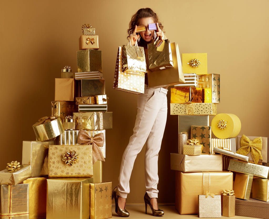 Ganzkörperporträt einer glücklichen jungen Frau in goldbeiger Hose und brauner Bluse mit Einkaufstaschen, die zwei Kreditkarten vor den Augen zwischen zwei Stapeln goldener Geschenke vor einer schlichten Wand halten. - Foto, Bild