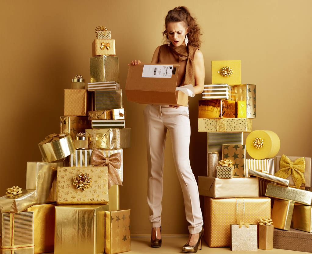 Retrato de larga duración de una joven sorprendida en pantalones beige dorado y blusa marrón entre 2 montones de regalos dorados mirando el paquete abierto. cliente decepcionado con la entrega o envío
. - Foto, imagen