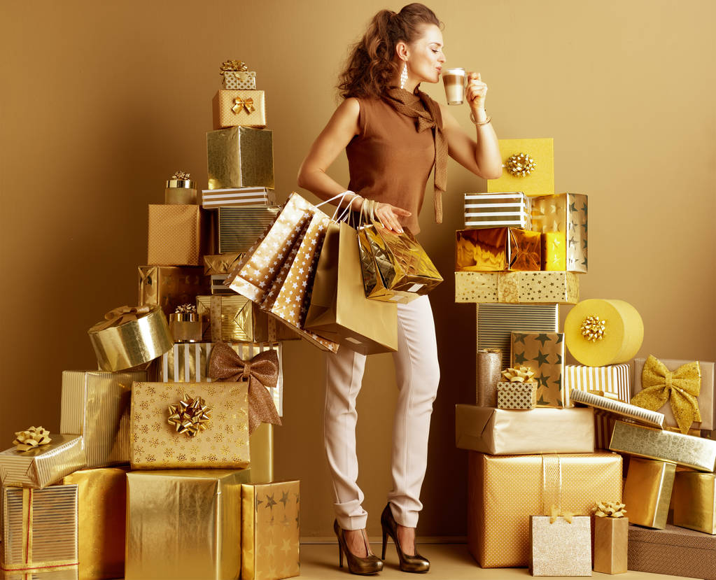 Retrato completo de moda-monger en pantalones beige oro y blusa marrón con portátil de oro bebiendo latte macchiato entre 2 pilas de regalos de oro delante de una pared lisa. compras libres de estrés
. - Foto, Imagen