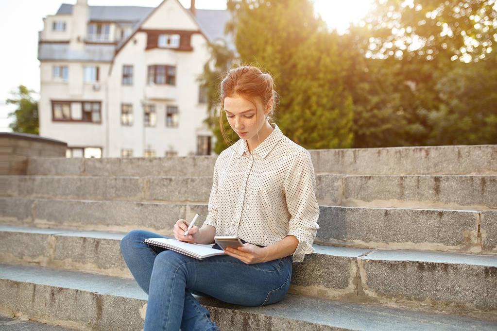 joven hermosa estudiante escribe un ensayo en su cuaderno sentado en las escaleras al aire libre Chica pelirroja con pecas estudio en tiempo libre
 - Foto, Imagen