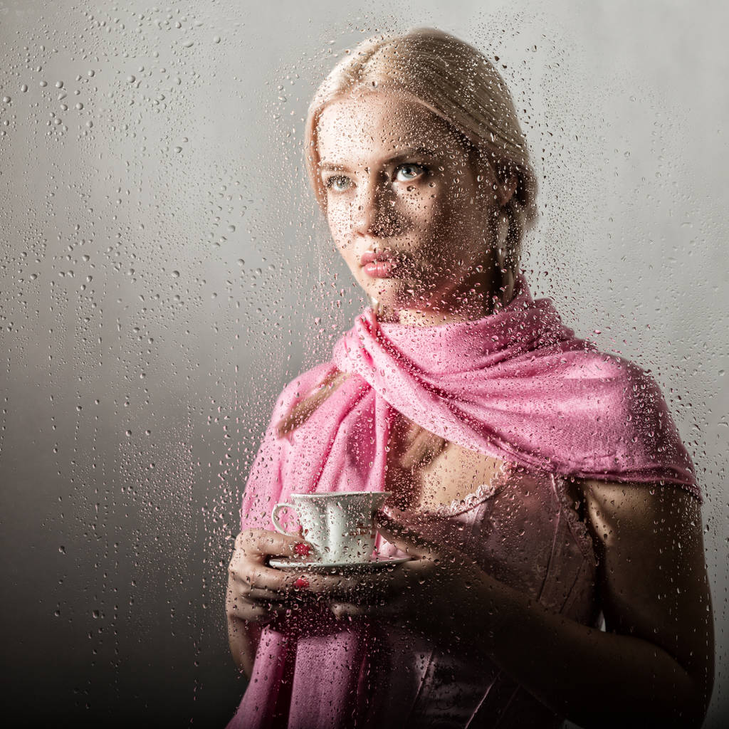молодая блондинка за стеклом с капельками воды. красивая девушка пьет кофе или чай
 - Фото, изображение