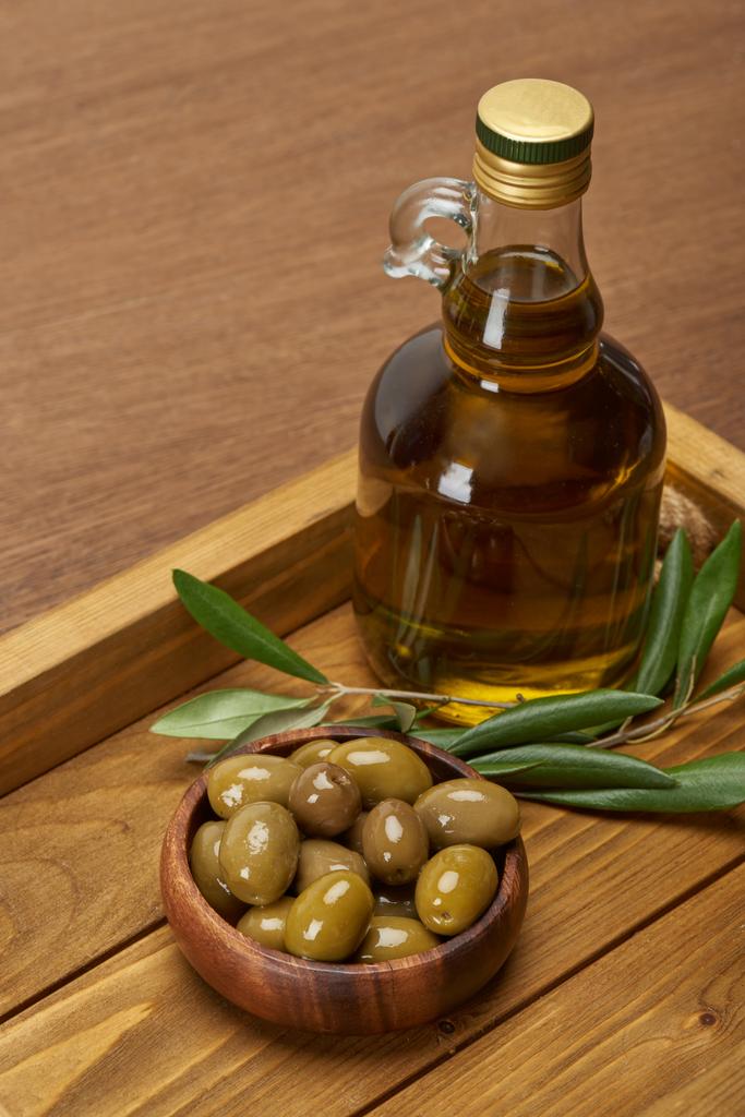 Dřevěný tác s olej láhev, misku oliv a olivových stromů listy na povrchu hnědá  - Fotografie, Obrázek