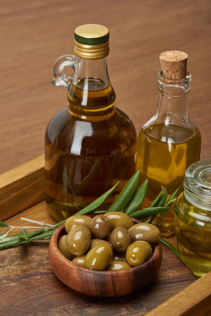 Holztablett mit verschiedenen Ölflaschen, Schüssel mit Oliven und Olivenblättern auf brauner Oberfläche  - Foto, Bild
