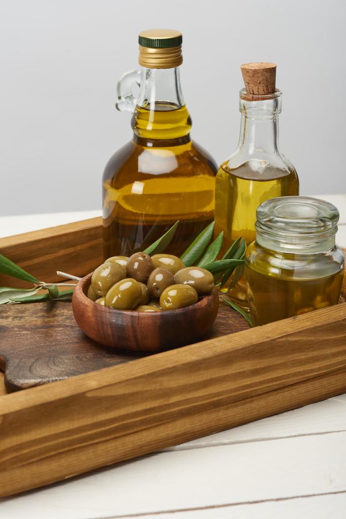 Holztablett mit einer Schüssel Oliven, Ölflaschen und Olivenbaumblättern auf weißer Oberfläche  - Foto, Bild
