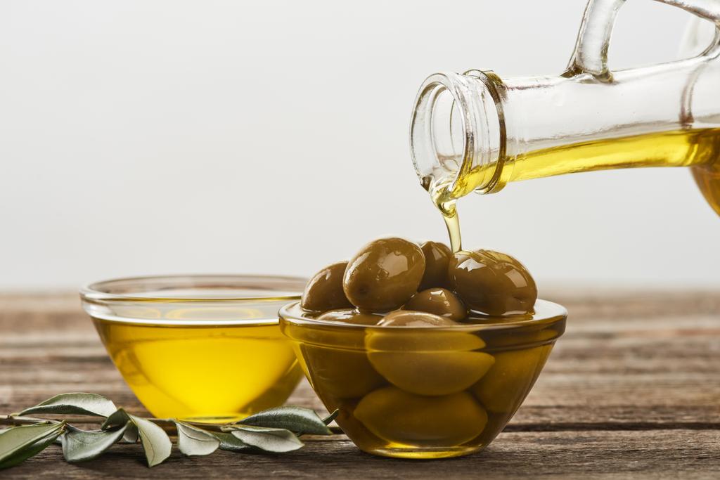 olie te gieten in een glazen kom met olijven, kuip vol olijven en olijfboom tak op houten oppervlak - Foto, afbeelding