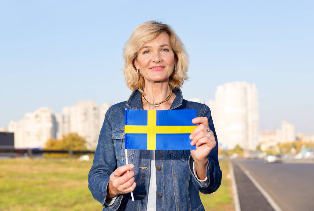 Mulher madura com a bandeira da Suécia contra o fundo da cidade e o céu azul
. - Foto, Imagem