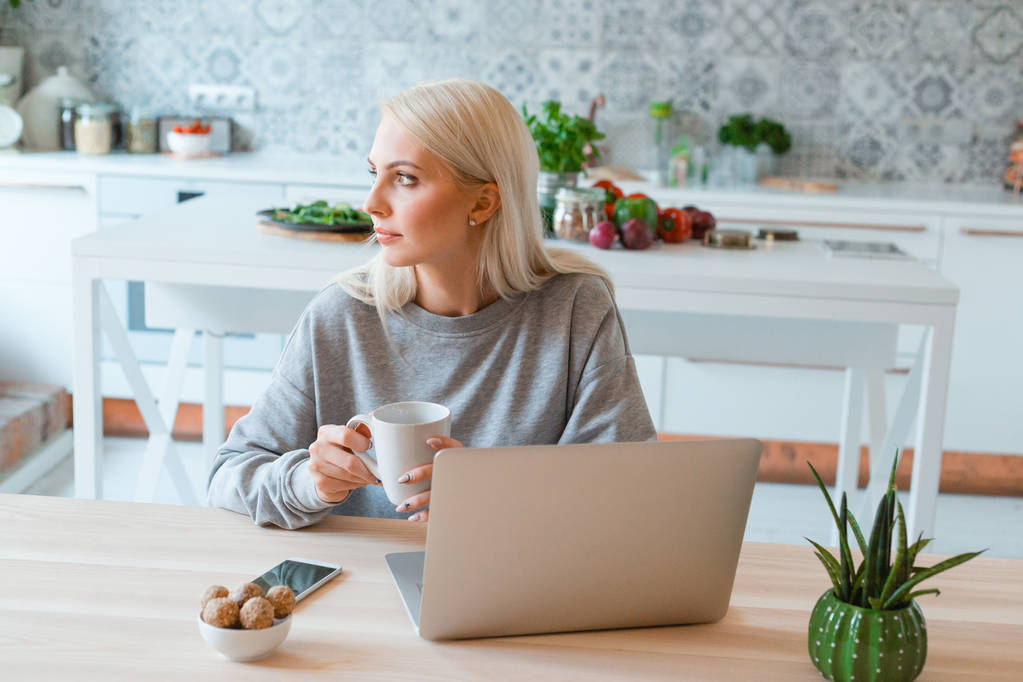 blonde junge Frau sitzt am Tisch in der Küche und trinkt Kaffee. Laptop auf dem Tisch.  - Foto, Bild