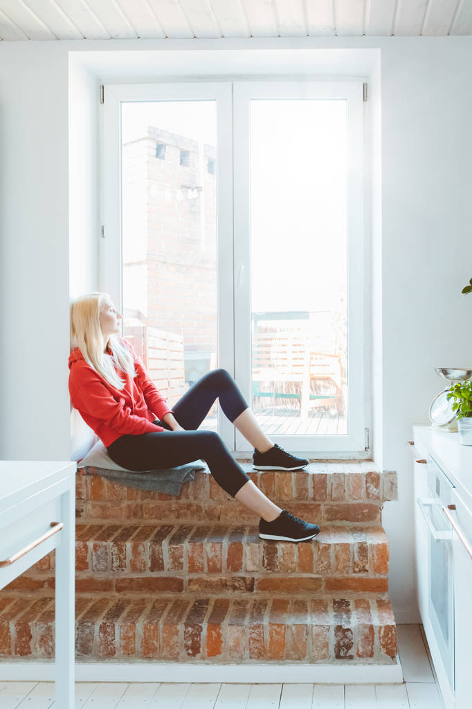 Mujer joven rubia con ropa deportiva descansando junto a la ventana en casa
.  - Foto, Imagen