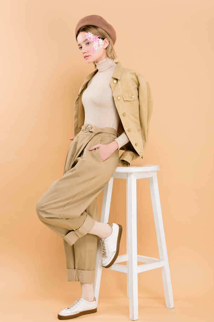 stylish woman in beret standing with hand in pocket near chair isolated on beige  - Photo, Image