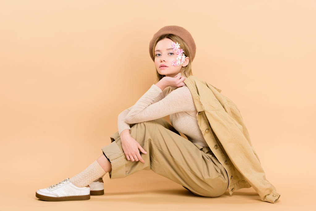 atraktivní dívka baret suport, zatímco sedí na podlaze izolované na béžové  - Fotografie, Obrázek