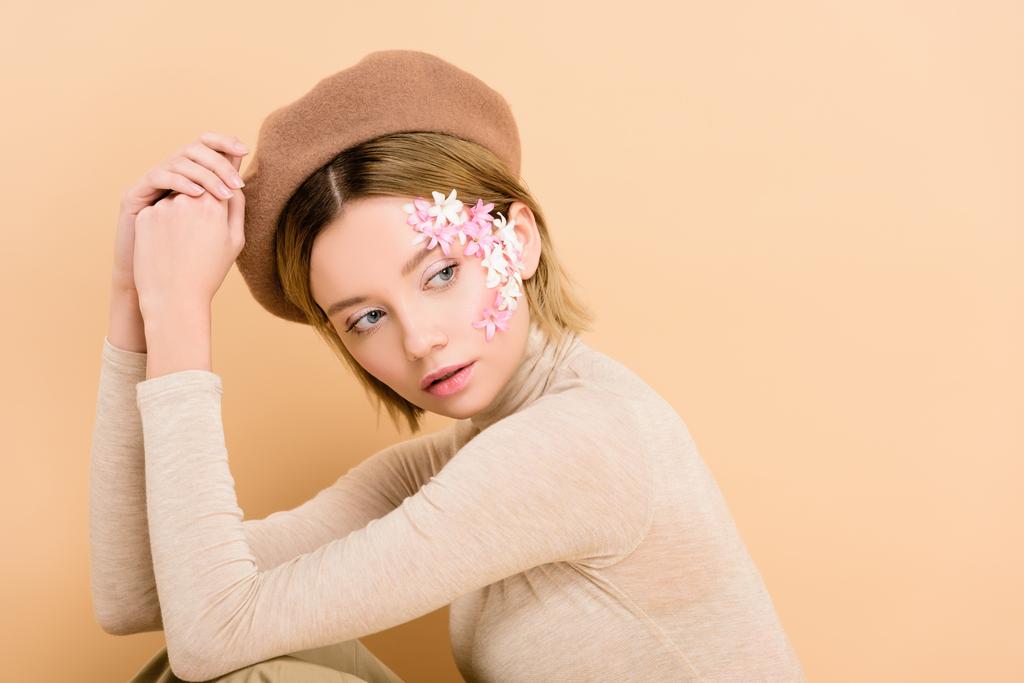 attraktive Frau mit Blumen im Gesicht trägt Baskenmütze isoliert auf beige  - Foto, Bild