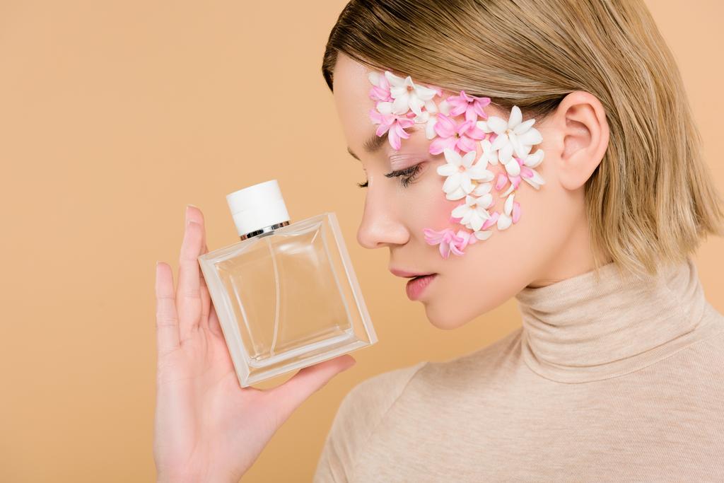 ελκυστική γυναίκα με λουλούδια στο πρόσωπο που ψάχνει στο μπουκάλι του αρώματος που απομονώνονται σε μπεζ - Φωτογραφία, εικόνα