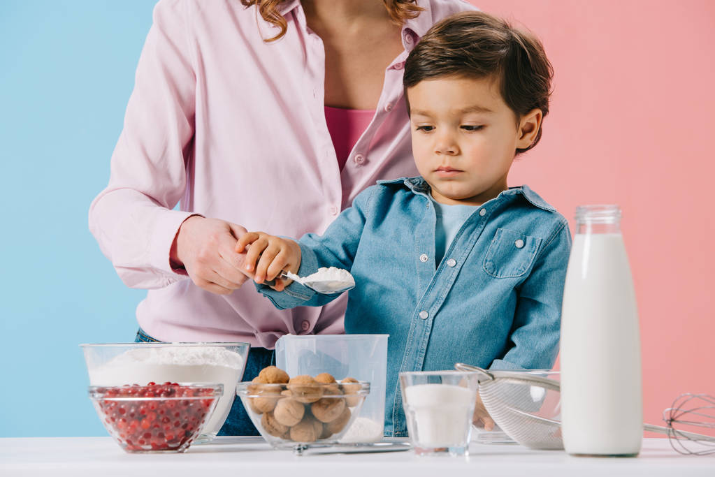 милый маленький мальчик с матерью наливая муку в измерительную чашку на двухцветном фоне
 - Фото, изображение