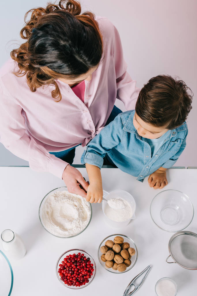top view of mother and little son pouring flour into measuring cup together on light background - Photo, Image