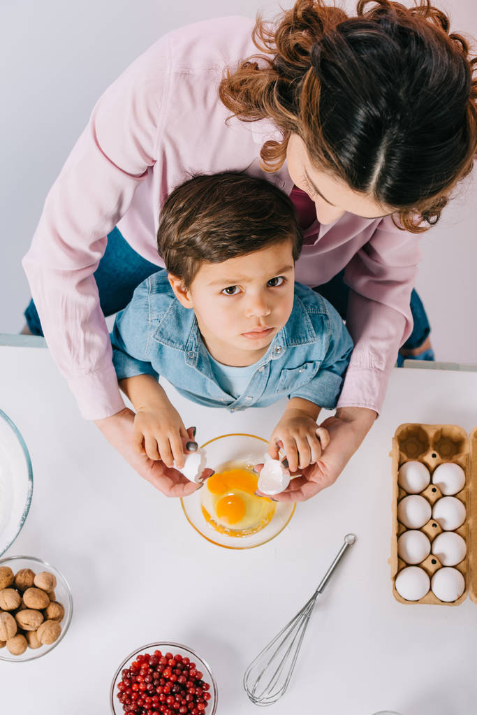 Draufsicht von Mutti mit ernstem kleinen Sohn, der Eier bricht, um Schüssel auf hellem Hintergrund - Foto, Bild