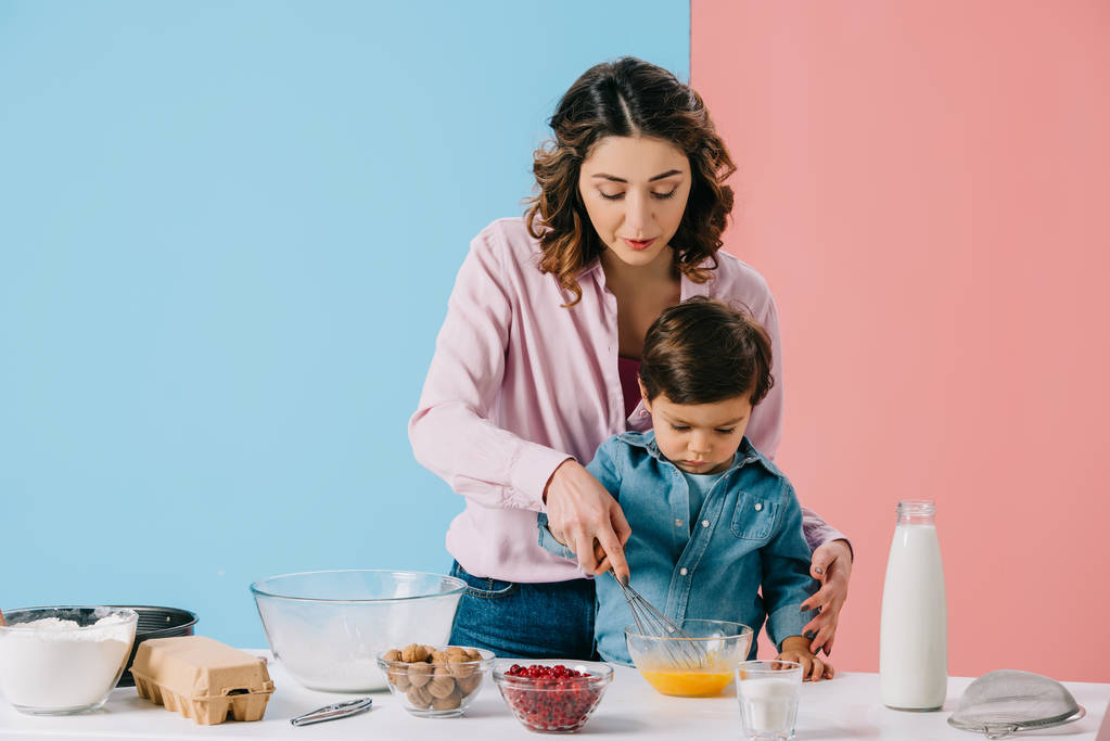 pretty mother with cute little son whipping eggs in bowl together on bicolor background - Photo, Image