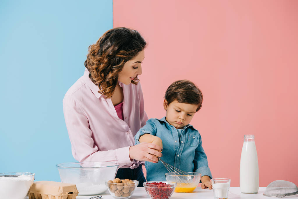 Мама с маленьким сыном взбивают яйца в миску, пока готовят вместе на двухцветном фоне
 - Фото, изображение