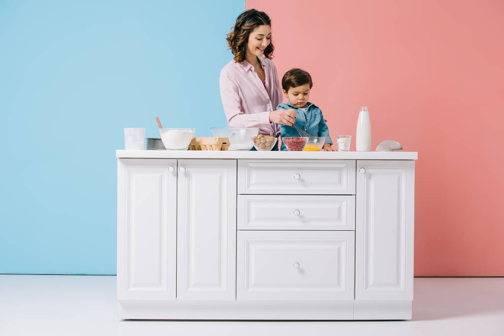 Gelukkig moeder met schattige zoontje koken samen op witte keukentafel op bicolor achtergrond - Foto, afbeelding