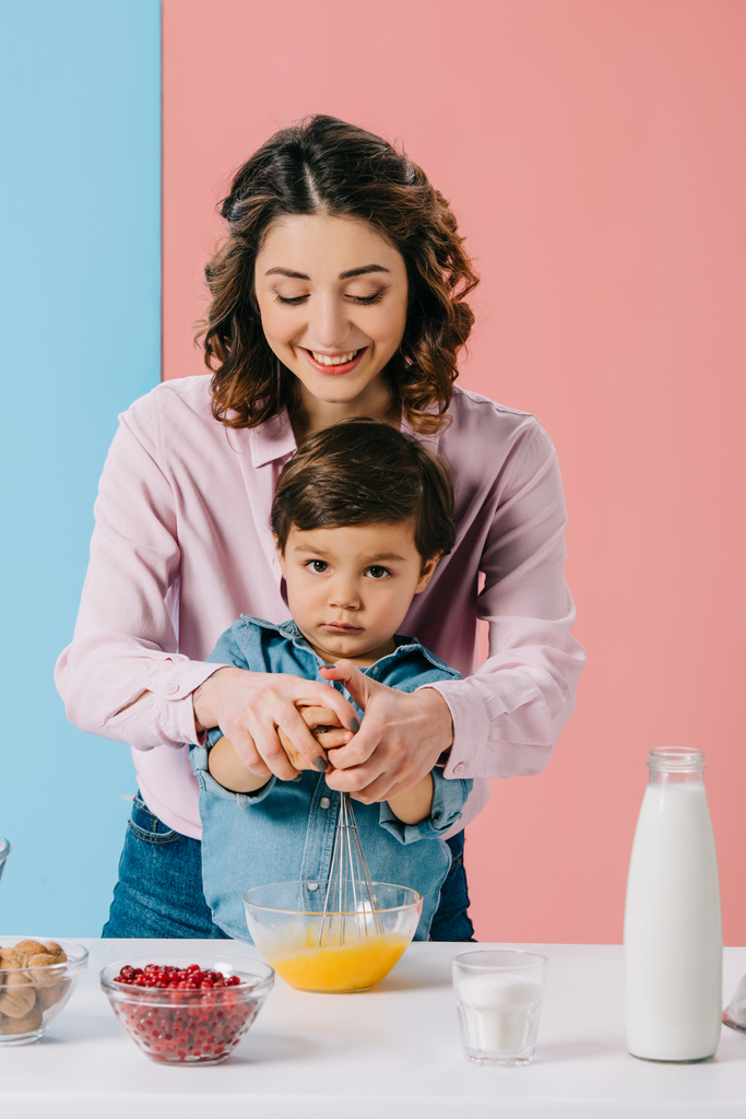 smiling mother with cute little son whipping eggs with balloon whisk while cooking together on bicolor background - Photo, Image