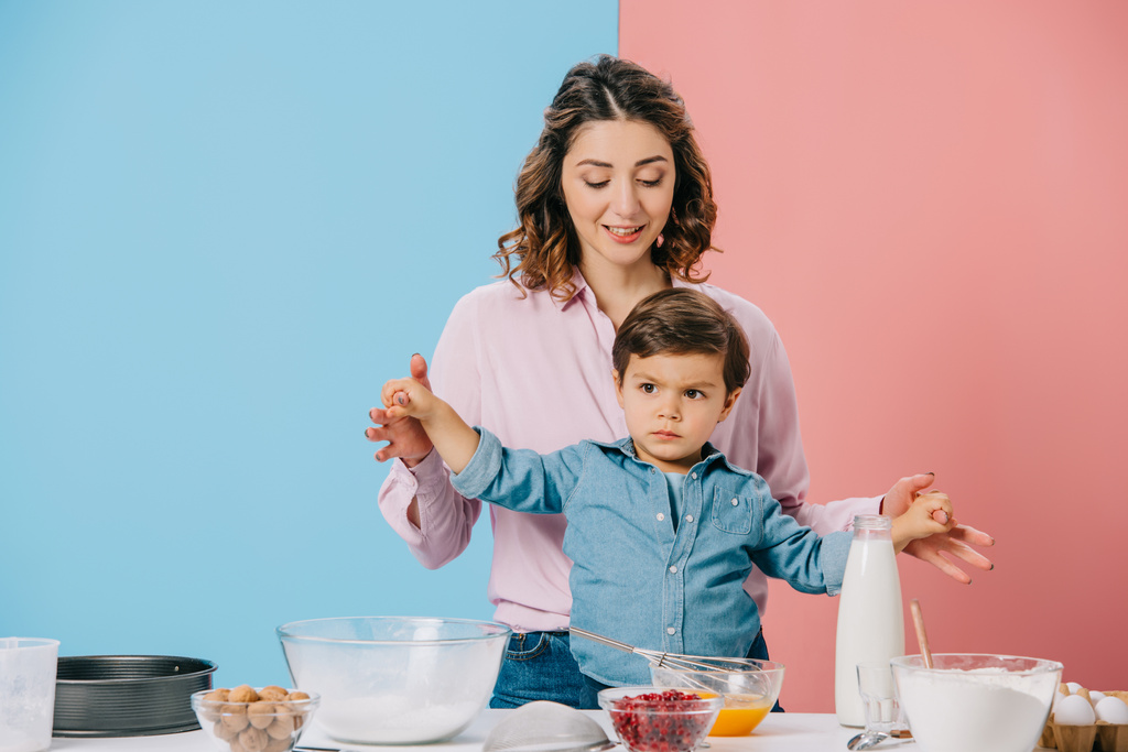 счастливая мать держит маленького сына за руки, стоя вместе на белом кухонном столе с кулинарным продуктом и посудой на двухцветном фоне
 - Фото, изображение