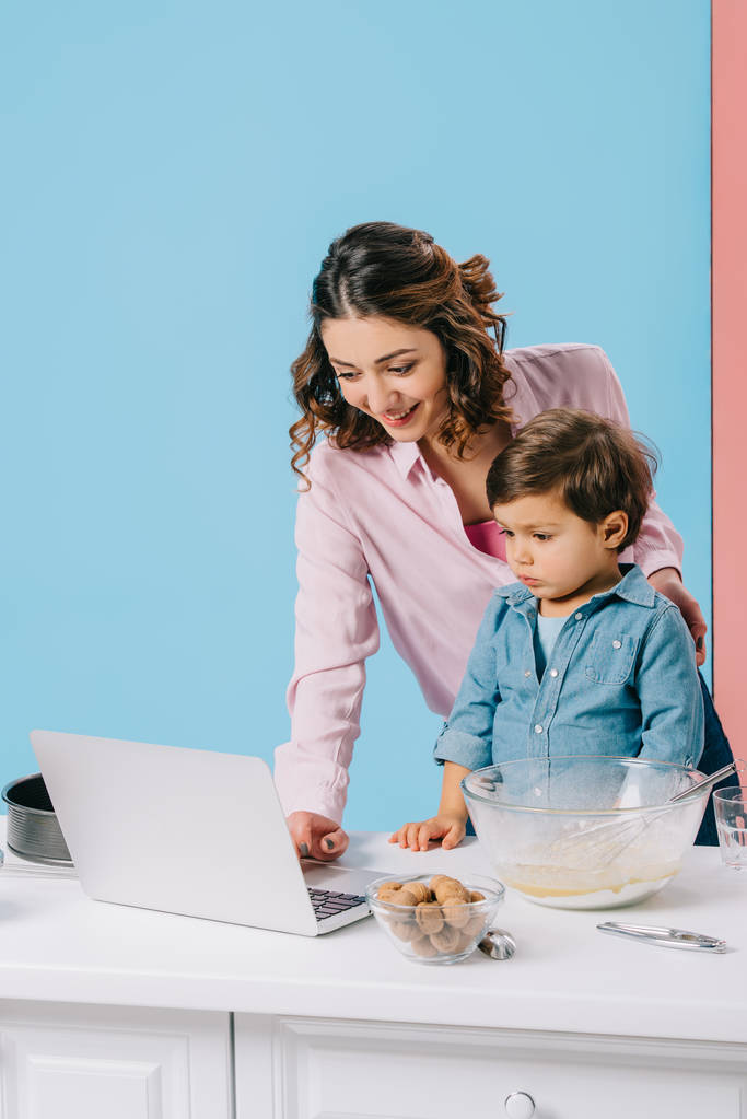 sonriente madre usando escritorio mientras cocina junto con su hijo pequeño sobre fondo bicolor
 - Foto, Imagen
