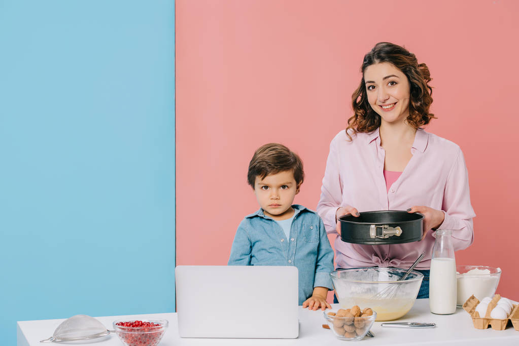 Ürünler, pişirme aletleri ve bicolor arka plan üstünde laptop ile mutfak masasında küçük oğlu ile birlikte yemek yaparken fırın formu tutan anne  - Fotoğraf, Görsel