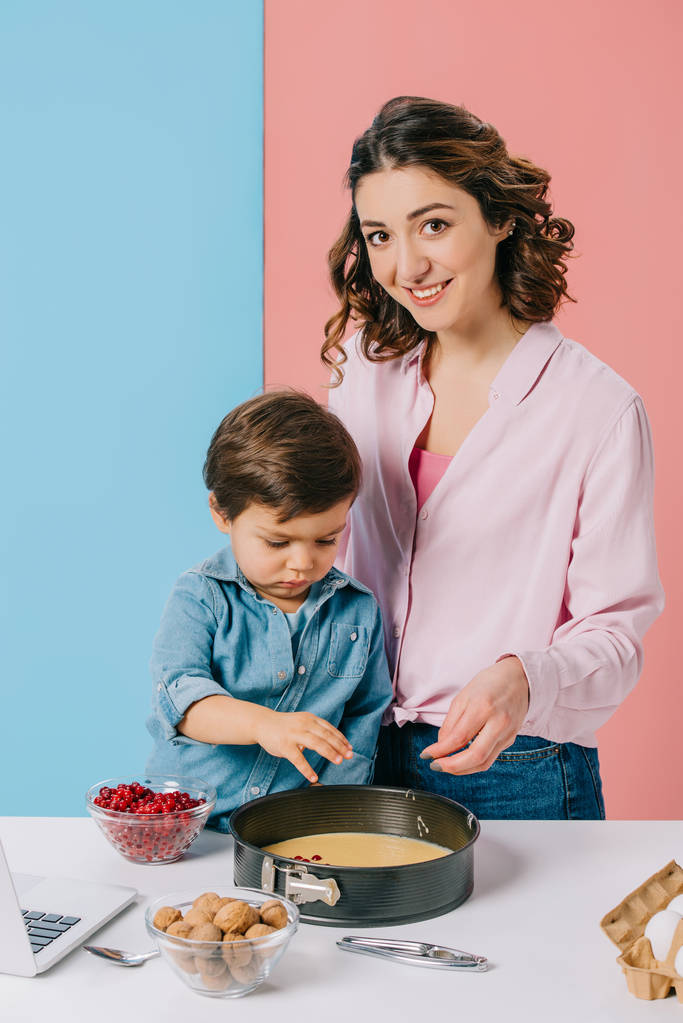Lächelnde Mutter mit entzückendem Sohn, der Preiselbeeren zum Teig in Backform auf zweifarbigem Hintergrund hinzufügt  - Foto, Bild