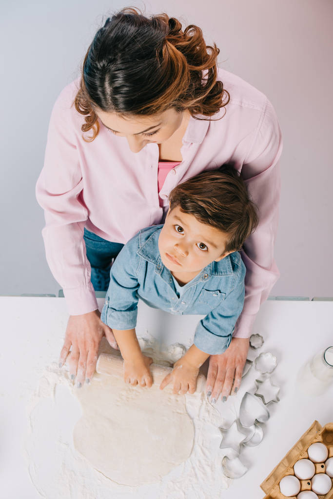 top view of mother with little son rolling out dough at kitchen table together on light background - Photo, Image
