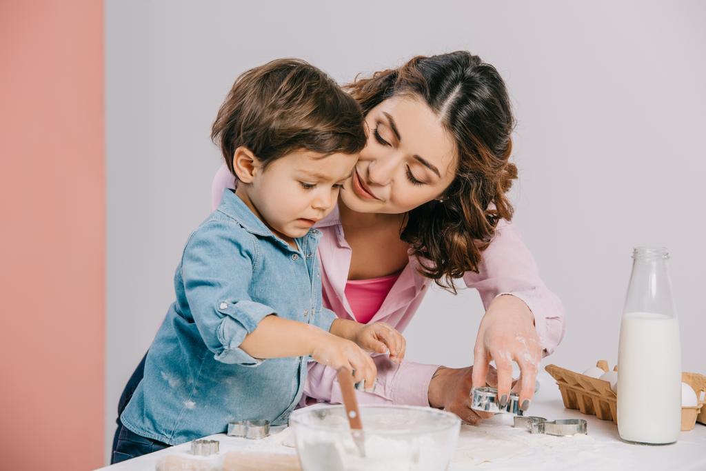 happy mother tenderly looking at cute little son while cooking pastry together on bicolor background  - Photo, Image