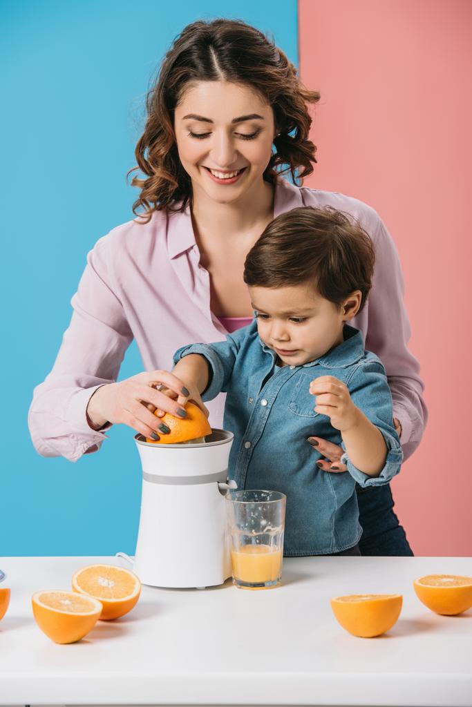 glückliche Mutter mit entzückendem Sohn, der zusammen frischen Orangensaft auf zweifarbigem Hintergrund presst  - Foto, Bild
