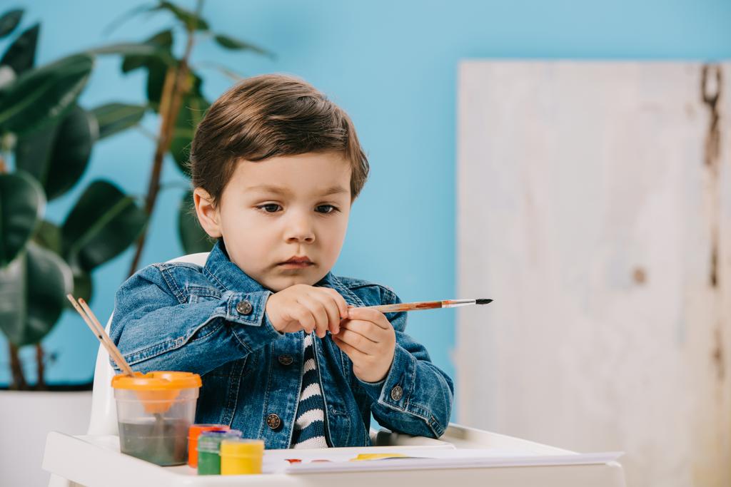 mignon petit garçon tenant pinceau de peinture tout en étant assis sur chaise haute avec des peintures à l'aquarelle sur la table
 - Photo, image