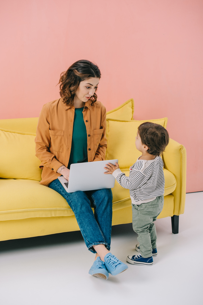 mother sitting on yellow sofa and using laptop while little son standing near - Photo, Image