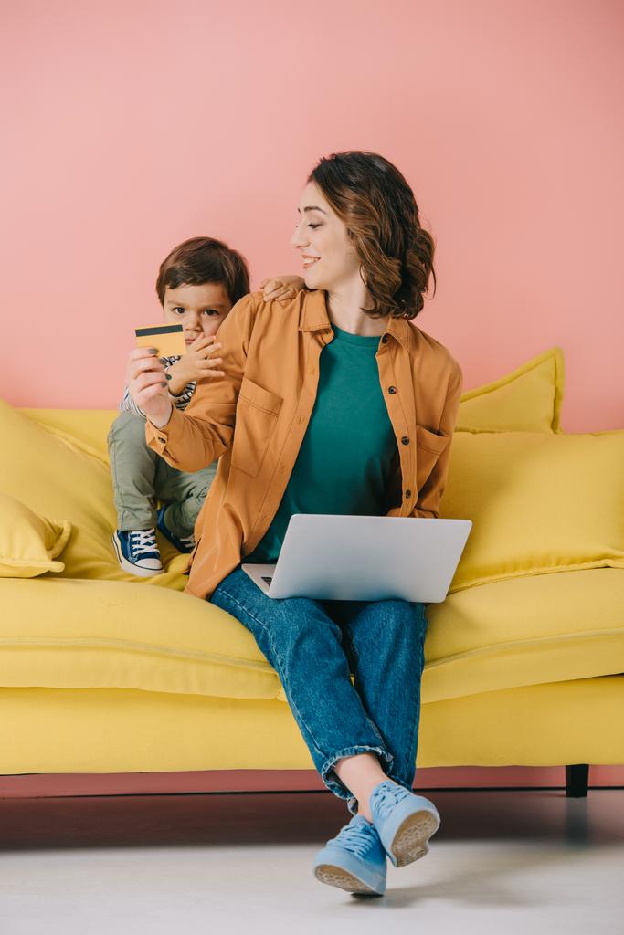 smiling mother showing credit card to little son while using laptop - Photo, Image