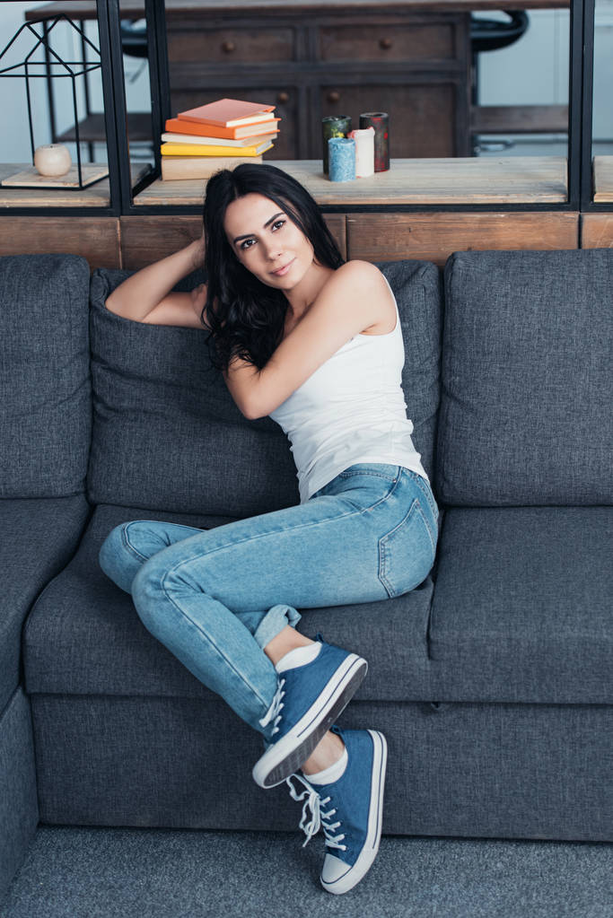 Hübsche Frau in Jeans sitzt auf Sofa und schaut in die Kamera - Foto, Bild
