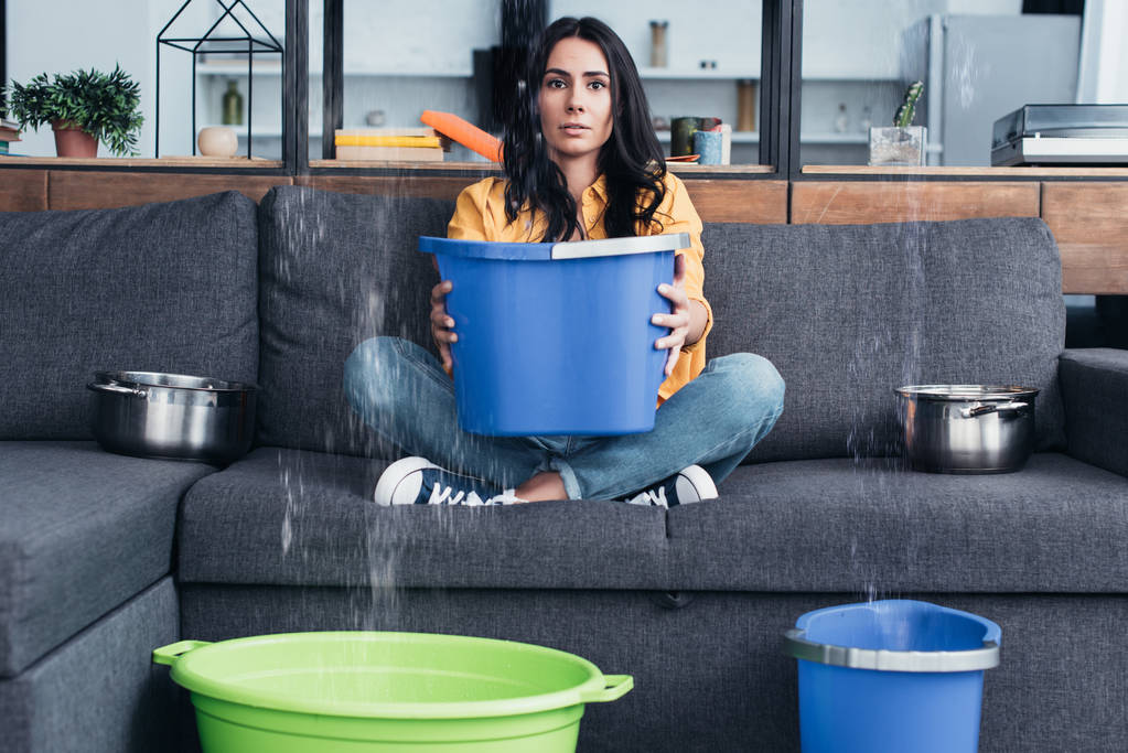 Bruneta žena držící kbelík na pohovce při poškození vodou v obývacím pokoji - Fotografie, Obrázek