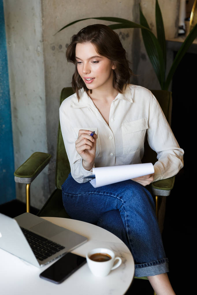 Image de jolie belle jeune femme assise dans un café à l'intérieur du travail avec ordinateur portable tenant des documents
. - Photo, image