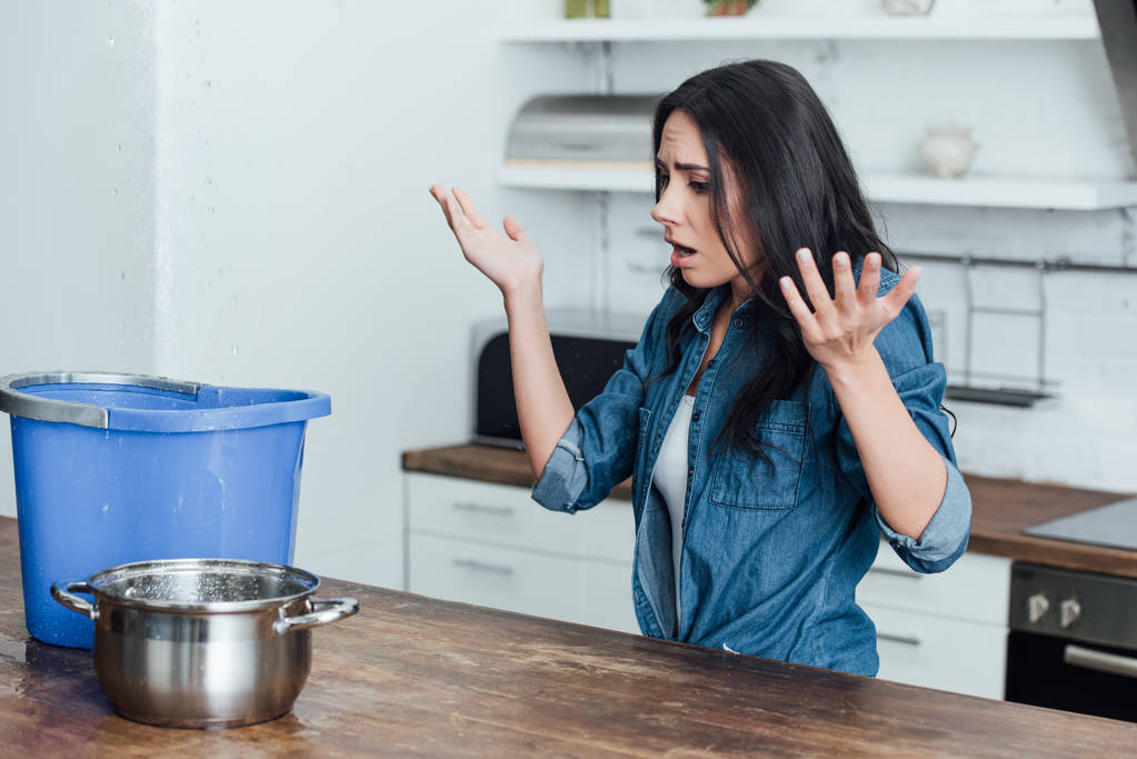 Грустная женщина с горшком и ведром размахивая руками во время протечки на кухне
 - Фото, изображение