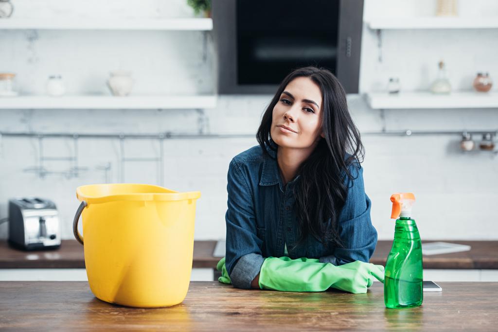 Beautiful brunette woman in rubber gloves sitting with bucket and spray in kitchen - Photo, Image