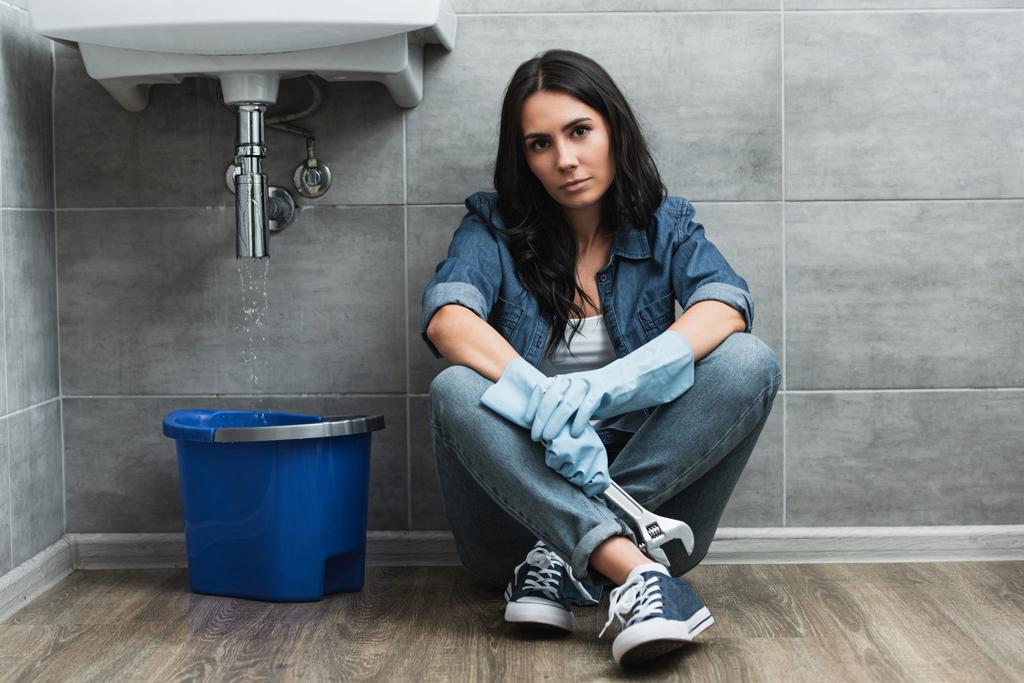Mujer con guantes de goma sentada junto al lavabo con llave inglesa
 - Foto, Imagen