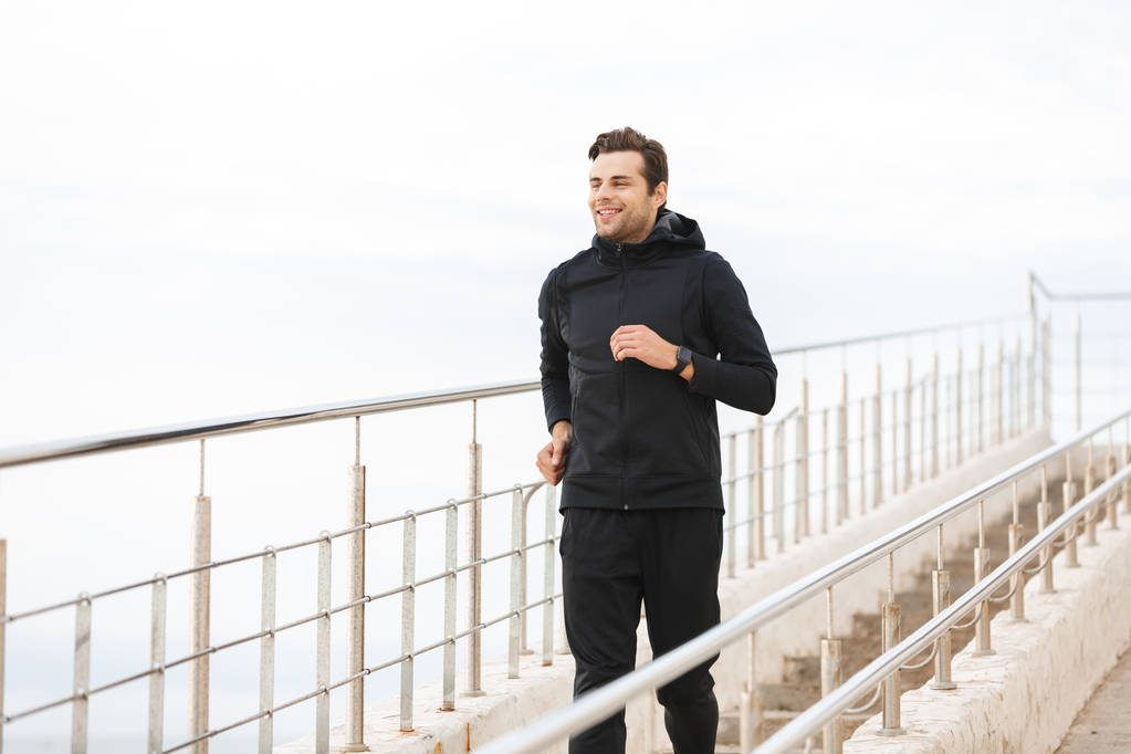 Image of satisfied sportsman 30s in black sportswear running along pier at seaside - Photo, Image